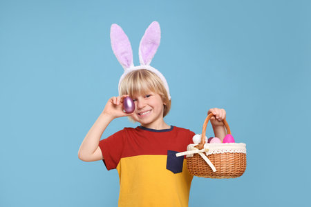 Happy boy in bunny ears headband holding wicker basket with painted Easter eggs on turquoise...