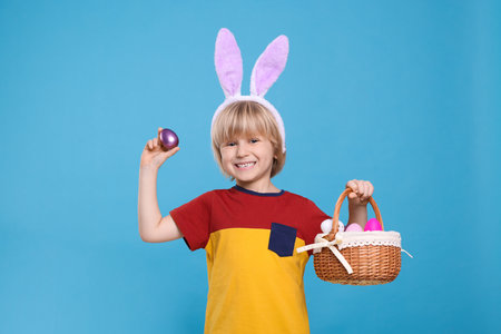Happy boy in bunny ears headband holding wicker basket with painted Easter eggs on turquoise...