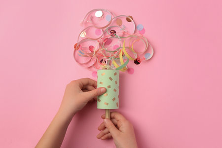 Woman holding party popper with serpentine and confetti on pink background, top view