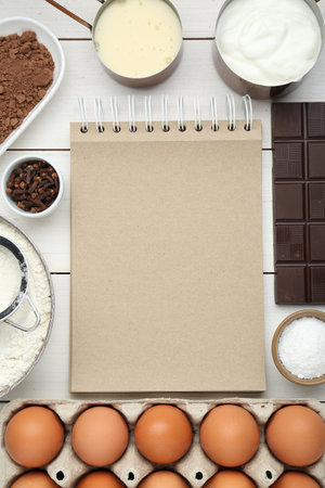 Blank recipe book surrounded by different ingredients on white wooden table, flat lay. Space for...