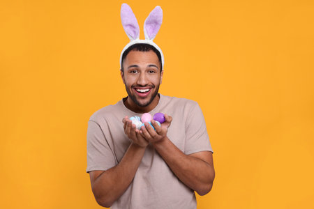 Happy African American man in bunny ears headband with Easter eggs on orange background