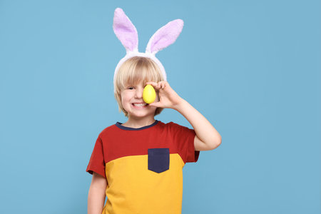 Happy boy in bunny ears headband holding painted Easter egg on turquoise background
