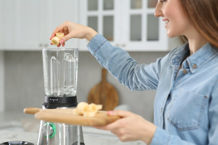 Beautiful young woman adding banana into blender for tasty smoothie in kitchen, closeupの写真素材