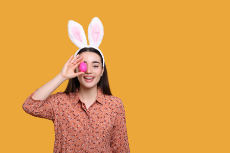 Happy woman in bunny ears headband holding painted Easter egg on orange background. Space for text