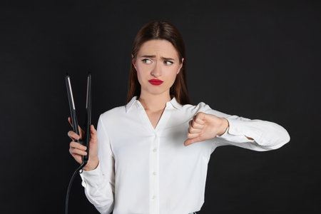 Upset young woman with flattening iron showing thumb down on black background. Hair damageの写真素材