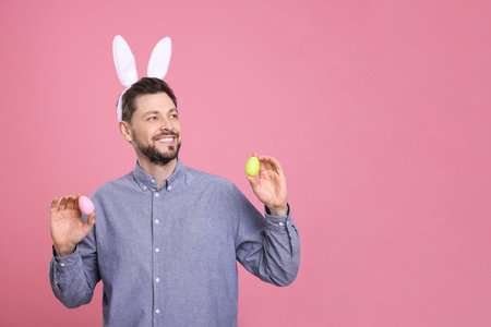Happy man in bunny ears headband holding painted Easter eggs on pink background. Space for textの写真素材
