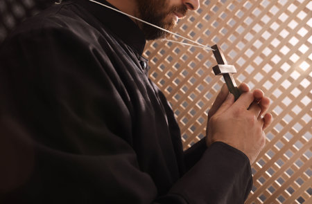Catholic priest in cassock holding cross in confessional booth, closeup