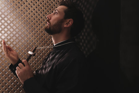 Catholic priest in cassock holding cross and praying to God in confessional