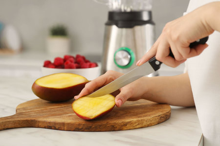 Woman preparing mango for tasty smoothie at white marble table in kitchen, closeup