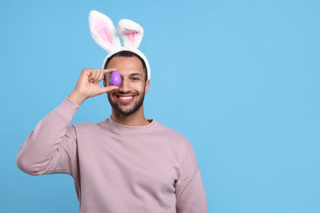 Happy African American man in bunny ears headband covering eye with Easter egg on light blue...