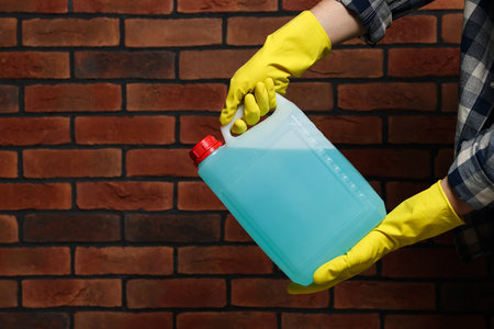Man in rubber gloves holding canister with blue liquid near brick wall, closeup. Space for textの写真素材