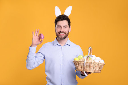 Portrait of happy man in cute bunny ears headband holding wicker basket with Easter eggs on orange backgroundの写真素材