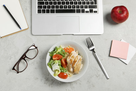 Bowl with tasty food, laptop, glasses, fork, apple and notebook on light gray table, flat lay. business lunchの写真素材