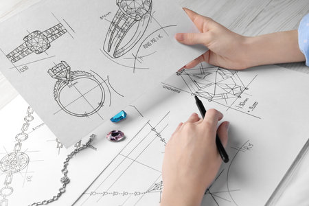 Jeweler with sketches of elegant jewelry at white wooden table, closeup
