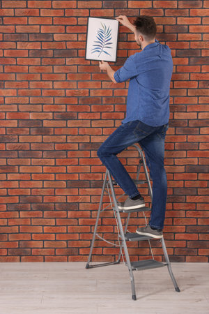 Man on stepladder hanging picture near red brick wallの写真素材