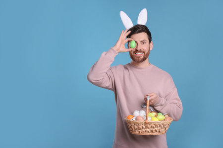 Happy man in cute bunny ears headband covering eye with Easter egg and holding wicker basket on...