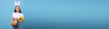 Happy woman with bunny ears holding bouquet and basket full of Easter eggs on light blue background, space for text. banner designの写真素材