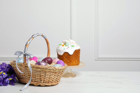 Easter basket with painted eggs near tasty cake and bouquet of flowers on white marble table. Space...