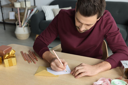 Man writing message in greeting card at wooden table in roomの写真素材
