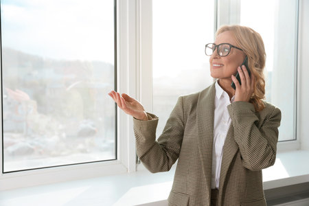 Lady boss talking on smartphone near window indoors, space for text. Successful businesswomanの写真素材