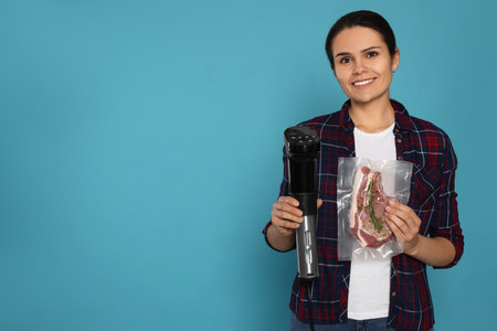 Beautiful young woman holding sous vide cooker and meat in vacuum pack on light blue background. Space for textの写真素材