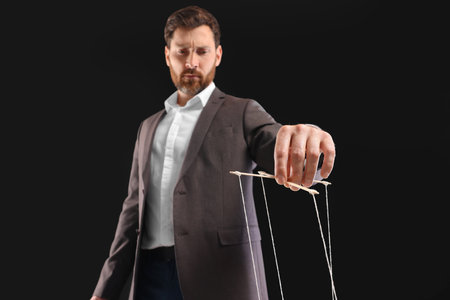 Man in suit pulling strings of puppet on black background, low angle viewの写真素材