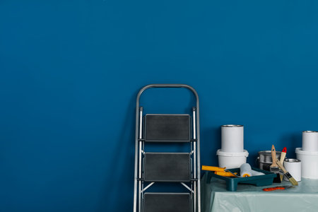 Metallic folding ladder and painting tools on table near blue wall, space for textの写真素材
