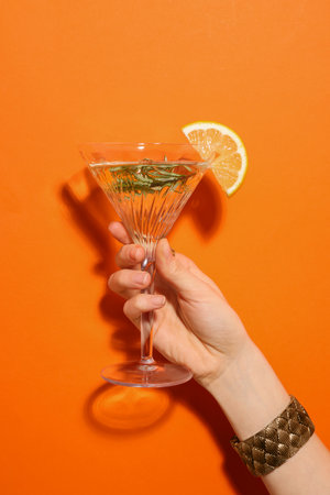 Woman holding martini glass of refreshing cocktail with lemon slice and rosemary on orange background, closeupの写真素材