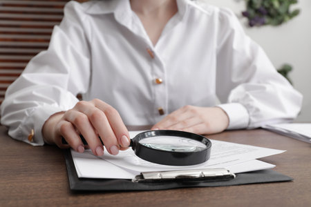 Woman looking at document through magnifier at wooden table indoors, closeup. Searching conceptの写真素材