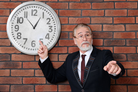 Angry senior boss pointing at clock on brick wallの写真素材
