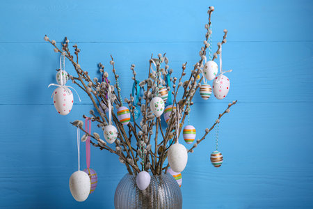 Beautiful willow branches with painted eggs in vase on light blue wooden background. Easter decor