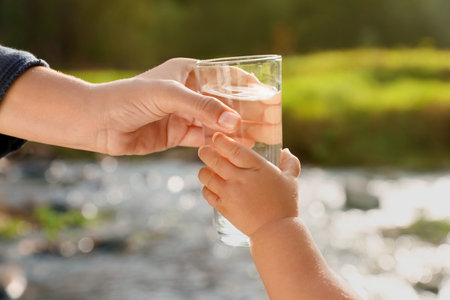 Mother giving her daughter glass of fresh water near stream on sunny day, closeupの写真素材