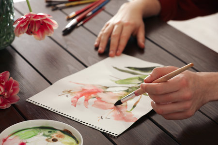 Woman painting flowers with watercolor at wooden table, closeup. Creative artworkの写真素材