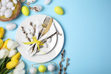 Festive table setting with tulips and painted eggs on light blue background, flat lay with space for...