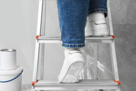 Woman climbing metallic folding ladder, can and bucket of paint near gray wall, closeup
