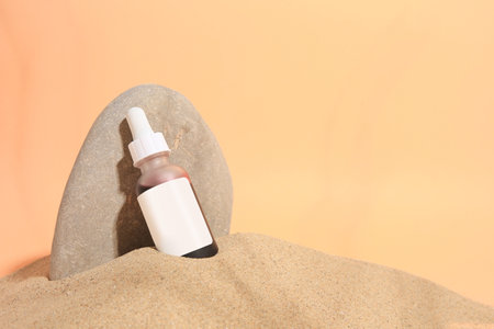 Bottle with serum and stone on sand against orange background, space for text. Cosmetic productの写真素材