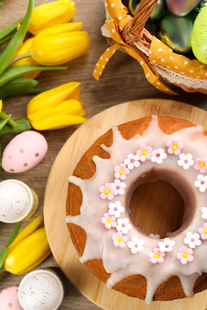 Delicious Easter cake decorated with sprinkles near painted eggs and tulips on wooden table, flat...