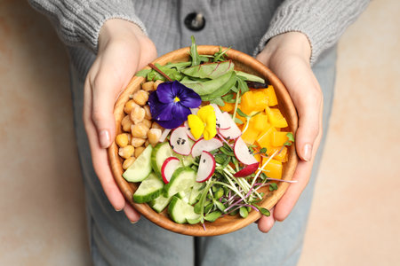 Woman holding delicious vegan bowl with cucumbers, chickpeas and violet flowers on beige background, closeupの写真素材