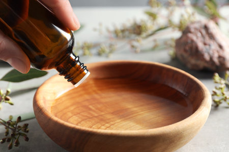 Woman dripping eucalyptus essential oil from bottle into bowl at light gray table, closeupの写真素材