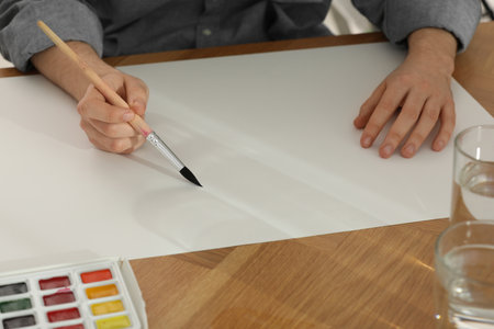 Man painting with watercolor on blank paper at wooden table, closeup
