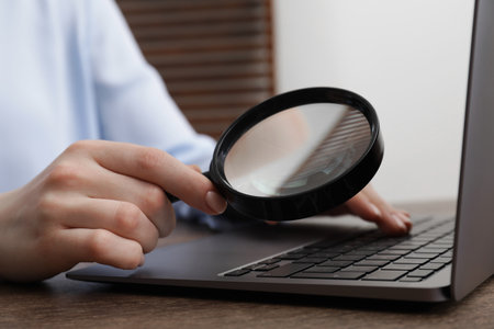 Woman holding magnifier near laptop at wooden table indoors, closeup. Online searching conceptの写真素材