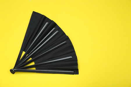 Stylish black hand fan on yellow background, top view. Space for text
