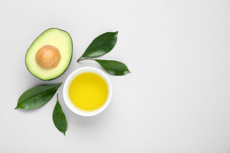 Bowl with oil, fresh cut avocado and leaves on white background, flat lay. Space for textの写真素材