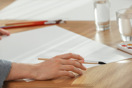 Man with brushes and blank papers at wooden table, closeup. Watercolor artwork