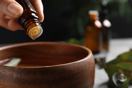 Woman dripping eucalyptus essential oil from bottle into bowl on blurred background, closeup. Space for textの写真素材