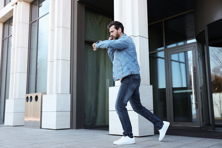 Emotional man talking on smartphone and looking at watch while running from building. Being late...
