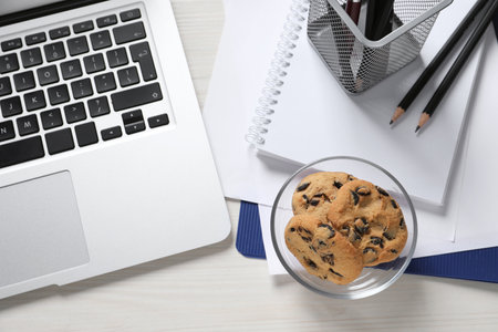 Bowl with chocolate chip cookies, laptop and office supplies on white wooden table, flat layの写真素材