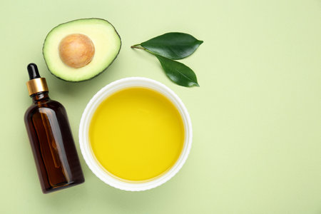 Essential oil, fresh cut avocado and leaves on light green background, flat lay. Space for text