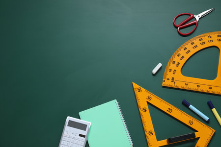 Flat lay composition with protractor, triangle and stationery on green chalkboard. Space for text