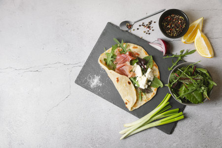 Delicious pita wrap with jamon, cheese cream and greens on light gray table, flat lay. Space for...